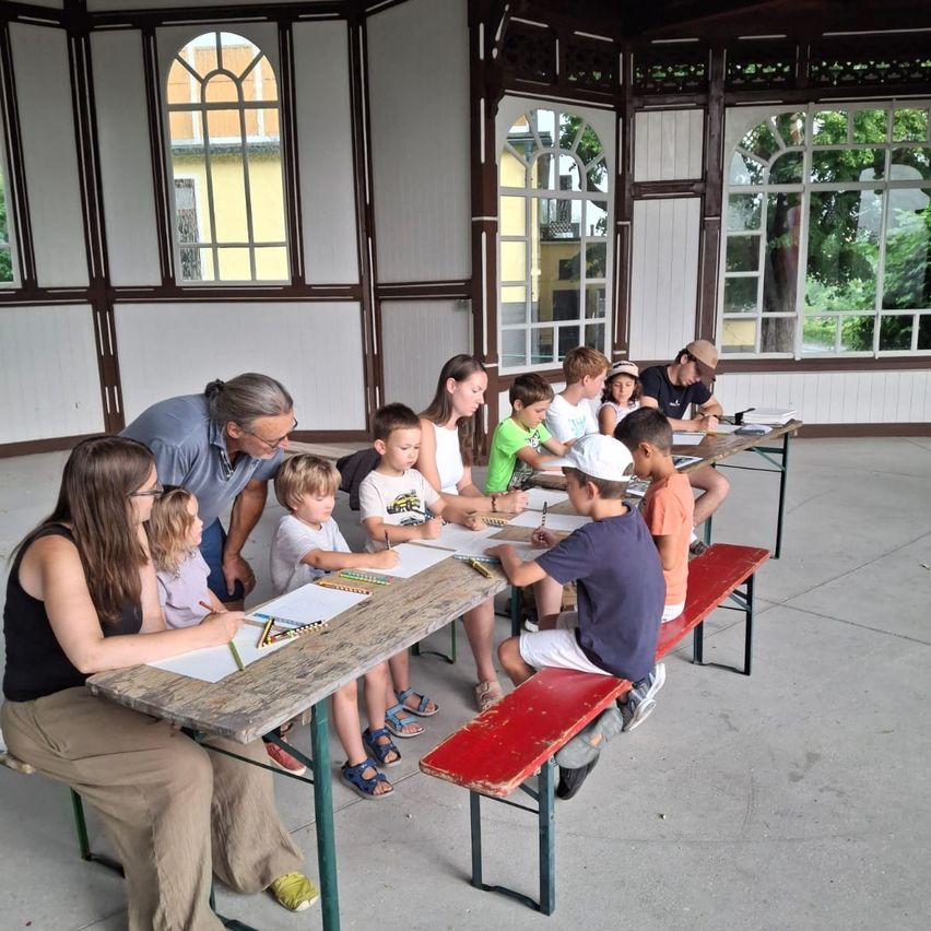 A group of children and adults are seated at tables in a gazebo, engaged in drawing activities. The gazebo has large windows and a yellow wall in the background.
