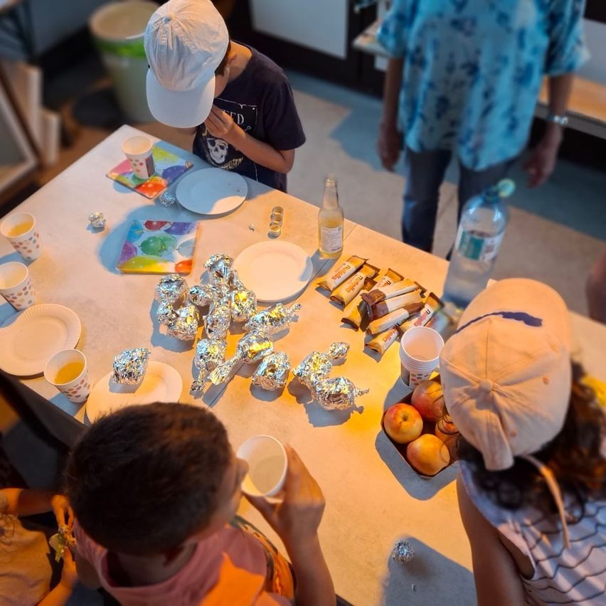 Children sit around a table with plates, cups, and snacks. One child eats while another looks at a wrapped food item. A person stands in the background.