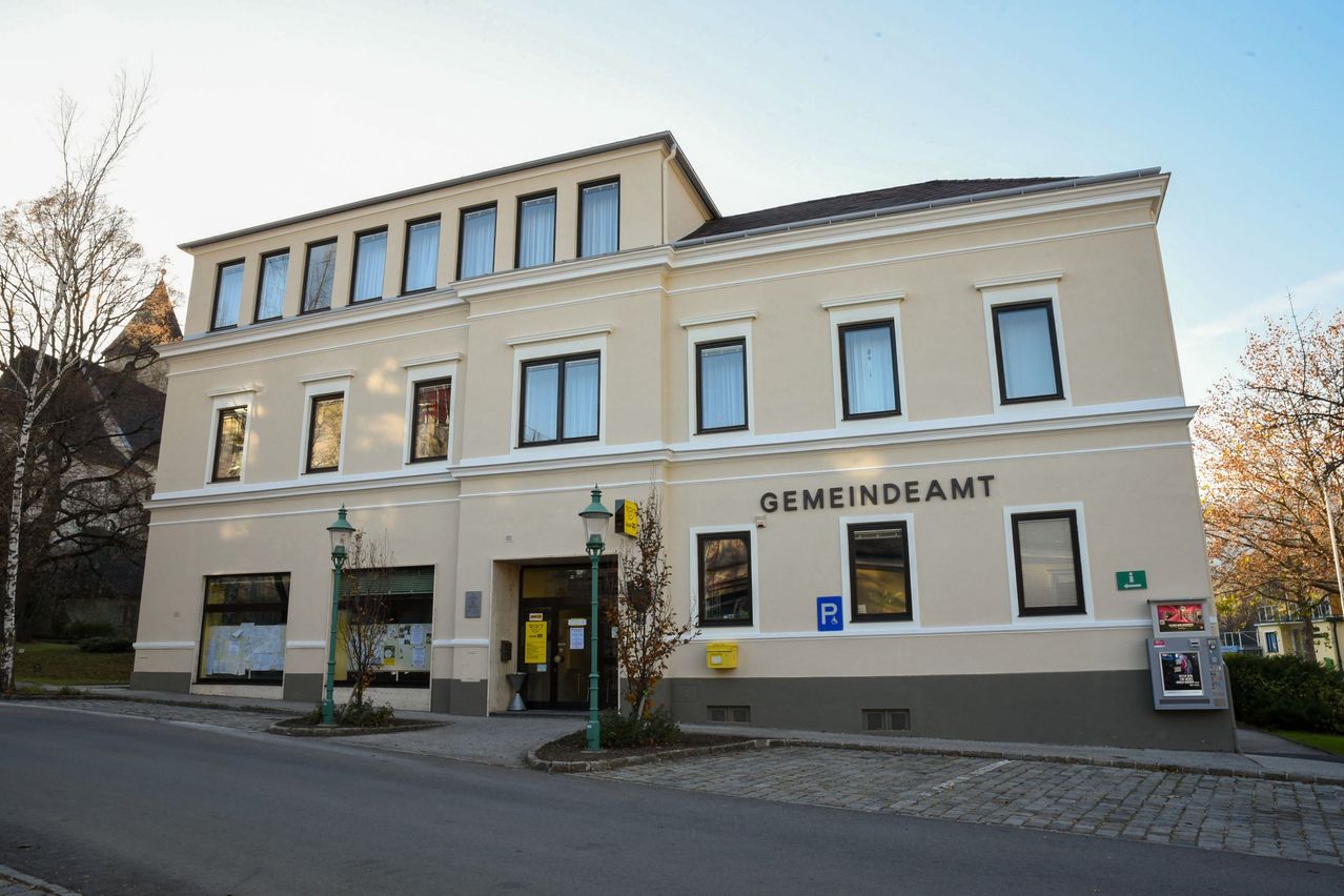 A beige building with multiple windows and a sign reading 'GEMEINDEAMT' above the entrance. Two green street lamps are in front.