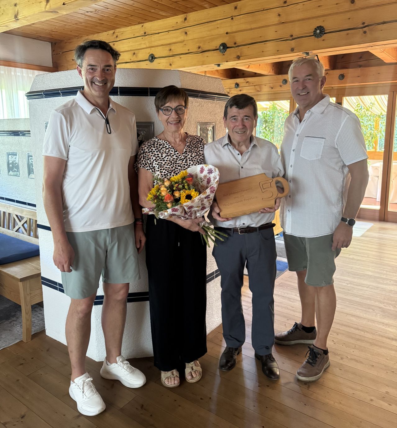 Vier Personen mit Blumenstrauß und Holztafel vor Kachelofen