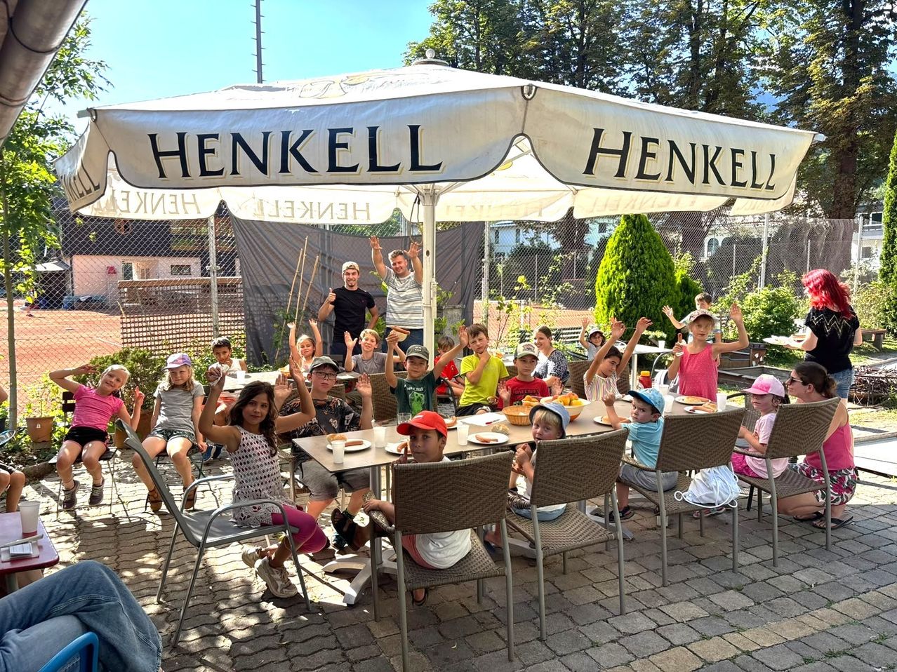 Eine Gruppe von Kindern und Erwachsenen sitzt unter einem Henkel-Schirm um Tische herum, hebt die Hände und lächelt.