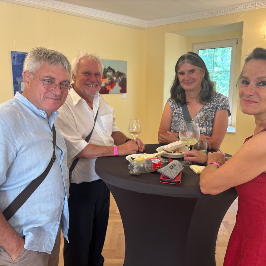 Vier Menschen lächeln und posieren für ein Foto bei einer Indoor-Veranstaltung. Zwei Männer stehen und zwei Frauen sitzen. Ein Tisch mit Weingläsern, Tellern und einer Flasche steht vor ihnen.