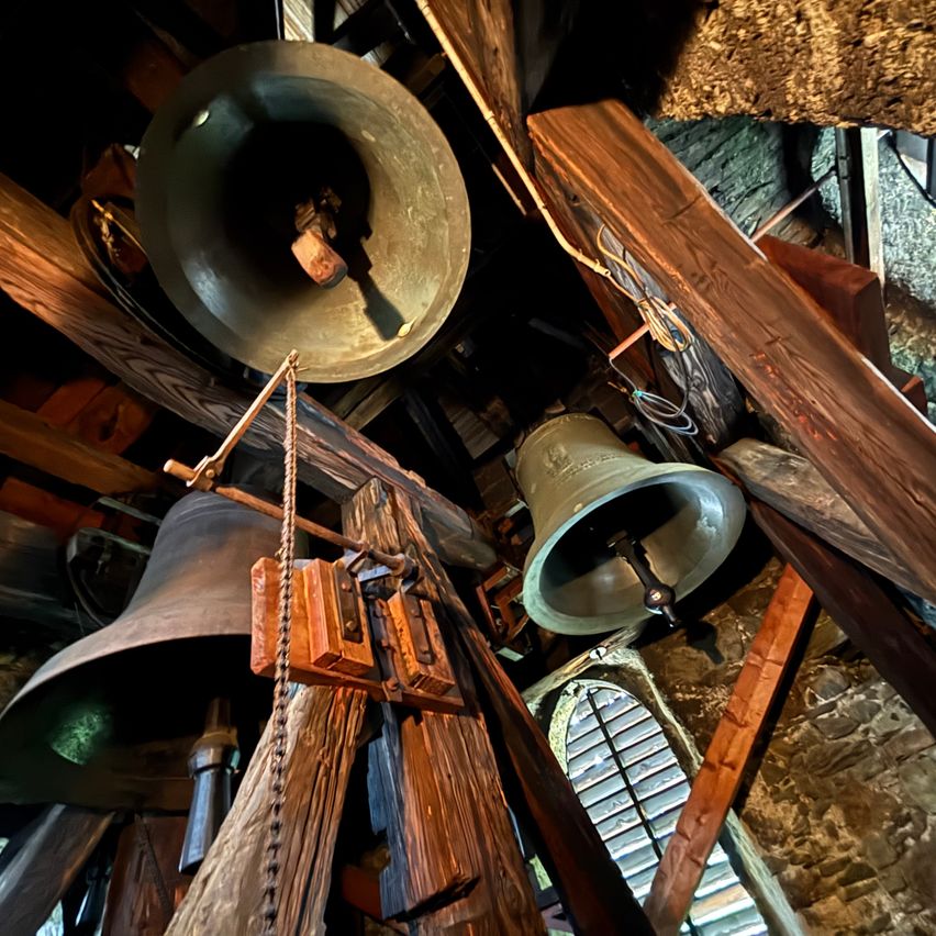 Drei große Glocken hängen von Holzbalken in einem Glockenturm, verbunden durch Seile und Holzmechanismen. Die Holzbalken und Steinmauern sind sichtbar.