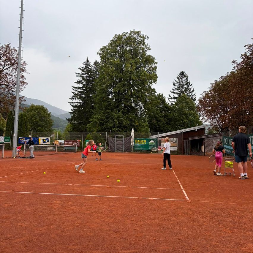 Eine Gruppe von Menschen spielt Tennis auf einem roten Sandplatz mit Bäumen und einem Gebäude im Hintergrund. Einige halten Tennisschläger und andere stehen in der Nähe eines Tennisballwagens.