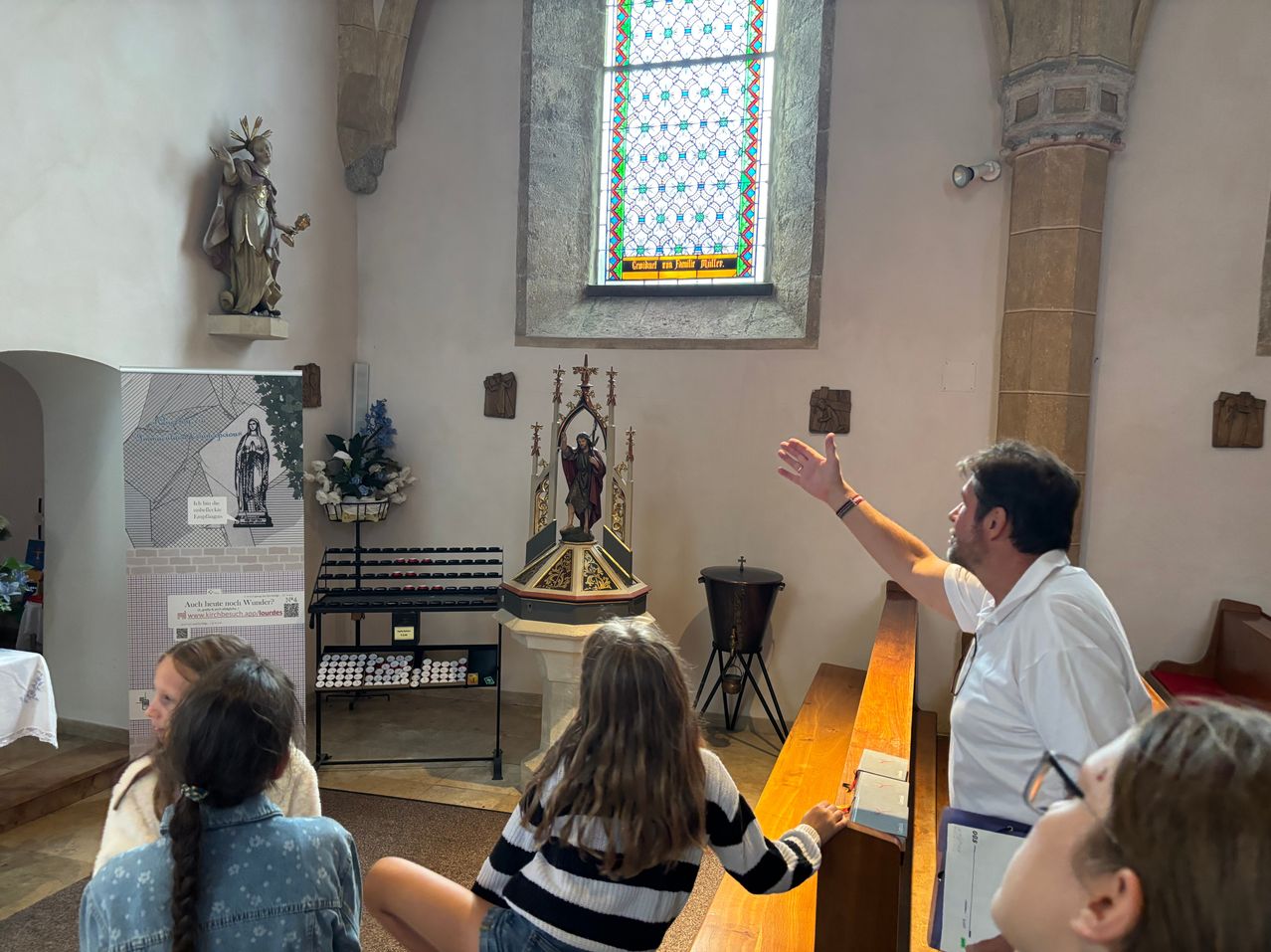 Ein Mann hält eine Präsentation in einer Kapelle mit einem Buntglasfenster, einer Statue und einem kleinen Altar. Mehrere Menschen sitzen und lauschen.