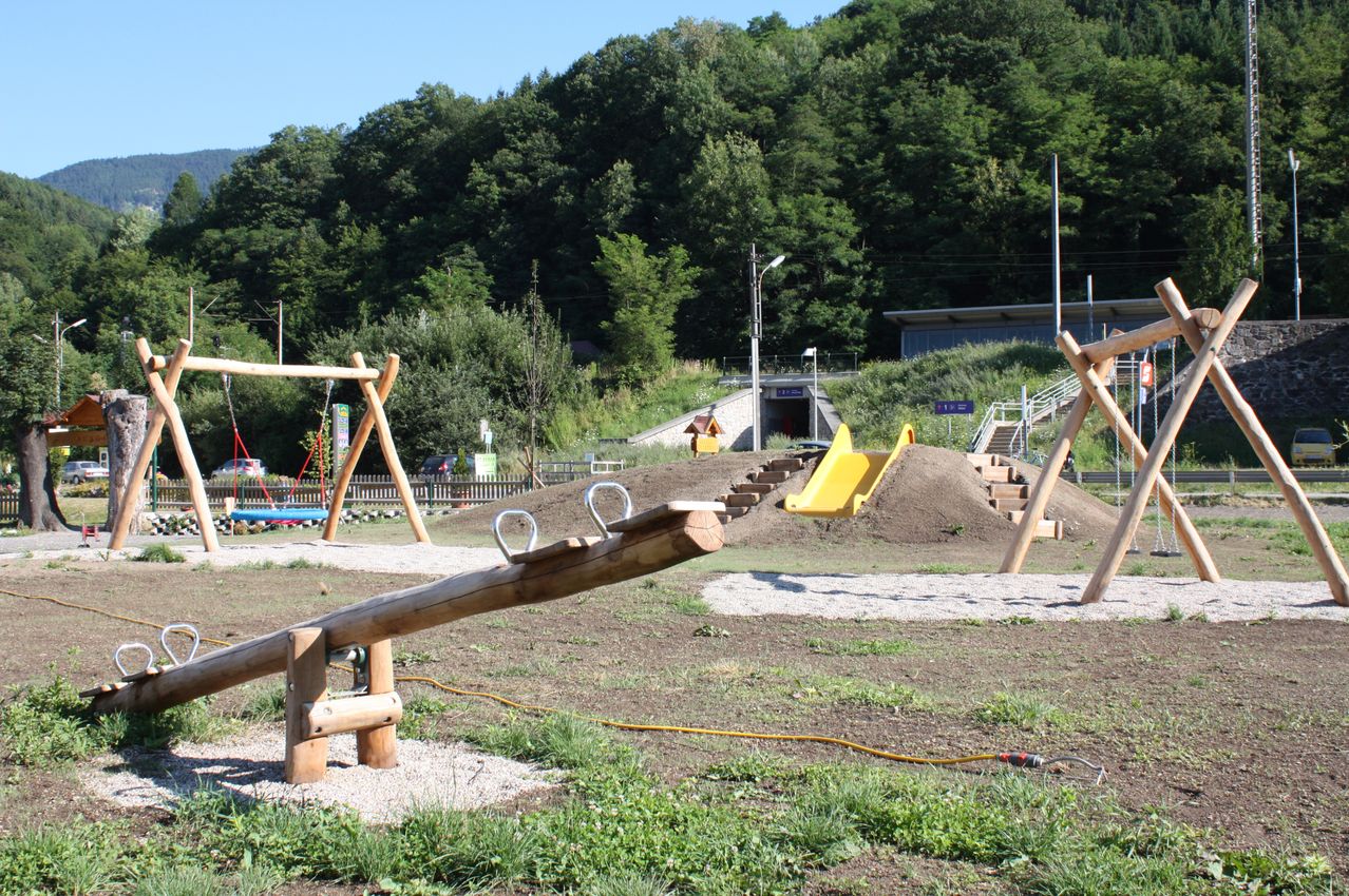An outdoor playground with wooden swings, a yellow slide, and a pile of dirt with steps. Trees and a building are in the background.