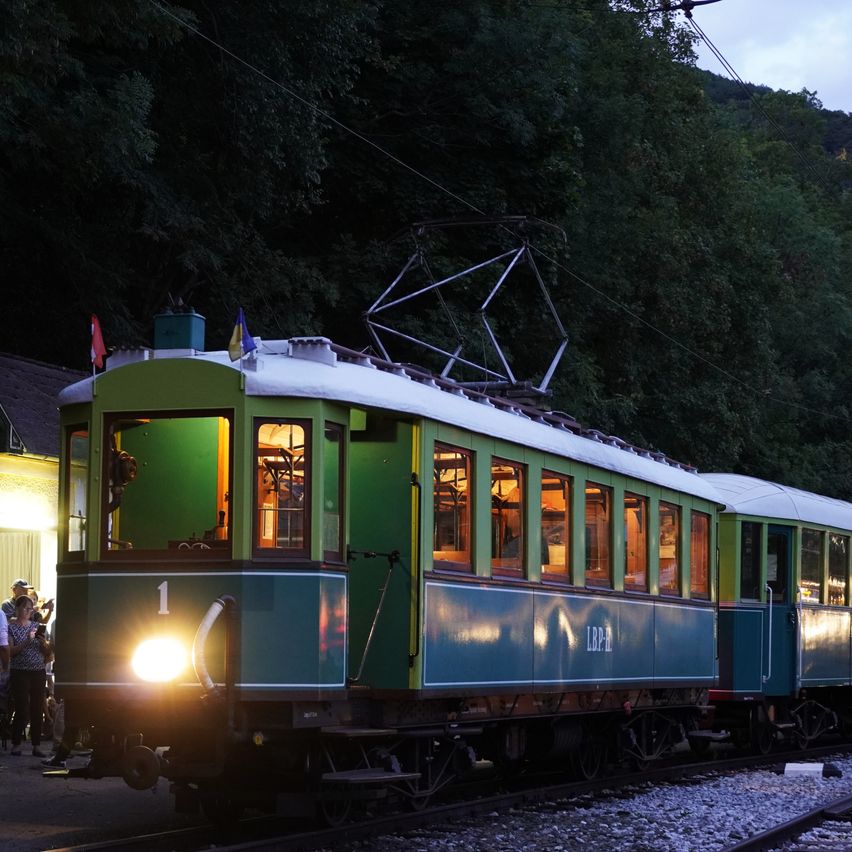 A vintage green train with the number 1 on the front is parked on a track, illuminated by its headlights. People are standing nearby, and flags are visible on the roof.