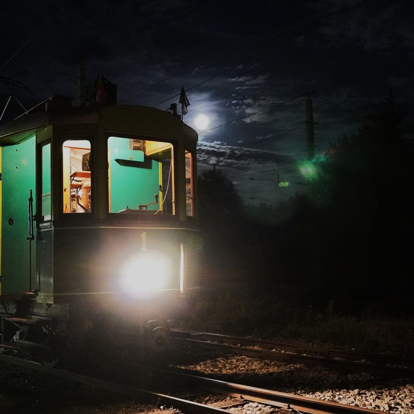 A vintage green tram is illuminated by its headlights as it travels on a railway track at night, with a full moon visible in the sky.