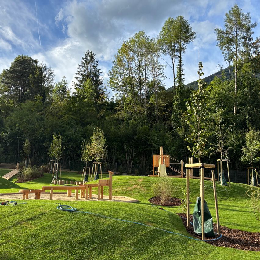 Ein gut gepflegter Außenspielplatz mit Holzstrukturen, Bänken und einer Rutsche, umgeben von üppigem Grün und Bäumen unter einem teilweise bewölkten Himmel.