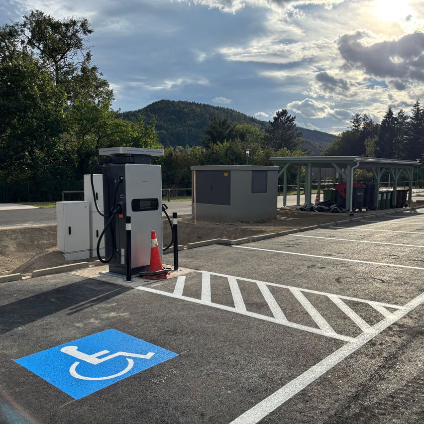 Eine Ladestation für Elektrofahrzeuge befindet sich auf einem Parkplatz, mit einem blauen Behindertenparkplatzschild und einem Kegel in der Nähe. Die Station ist von Bäumen und Bergen in der Ferne umgeben.