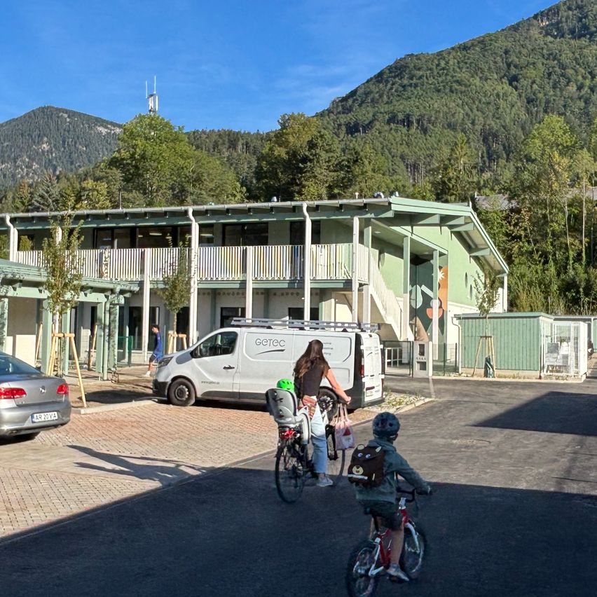 Eine Familie fährt Fahrräder vor einem modernen Gebäude mit Berghintergrund. Ein Van ist in der Nähe geparkt.