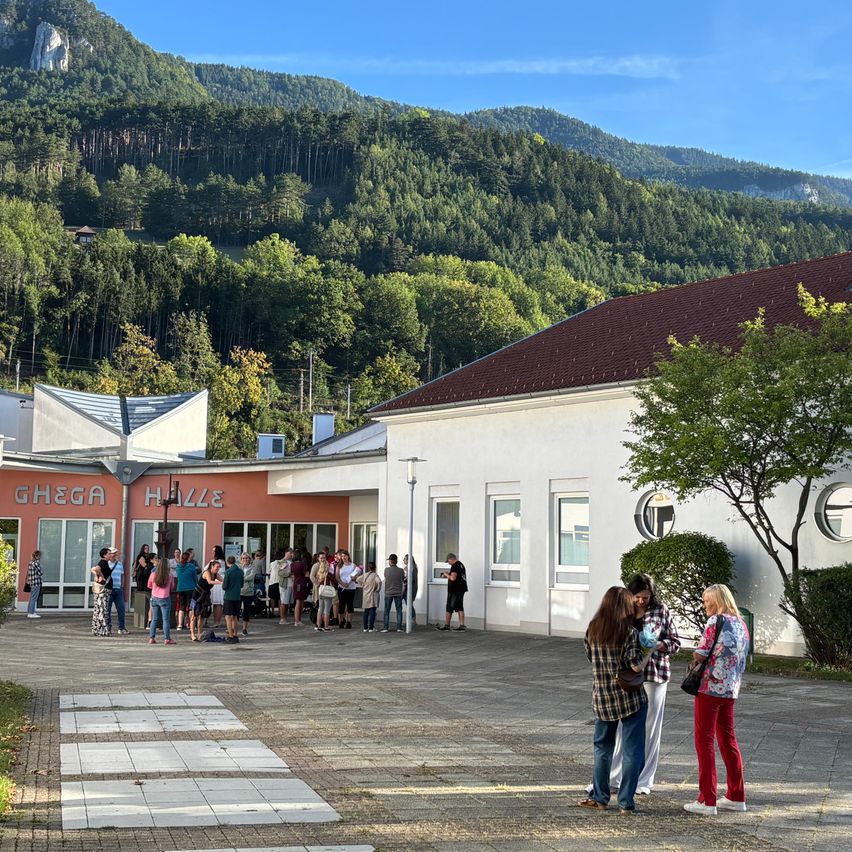 Eine Gruppe von Menschen steht in einer Schlange vor einem Gebäude mit einem roten Dach, umgeben von Bäumen und Bergen.