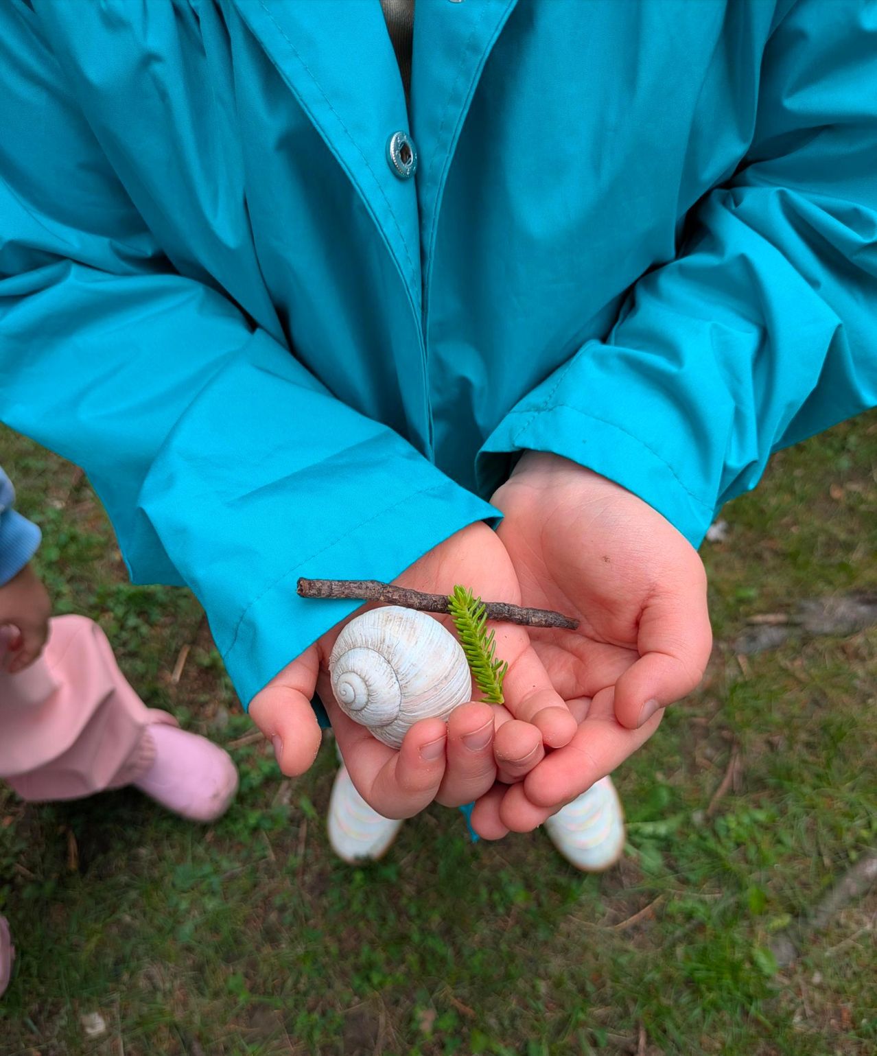 Ein Kind in einem blauen Regenmantel hält eine Schnecke, einen Zweig und eine grüne Sprossen auf einem Grasfeld. Ein weiteres Kind in rosa Hose steht in der Nähe.