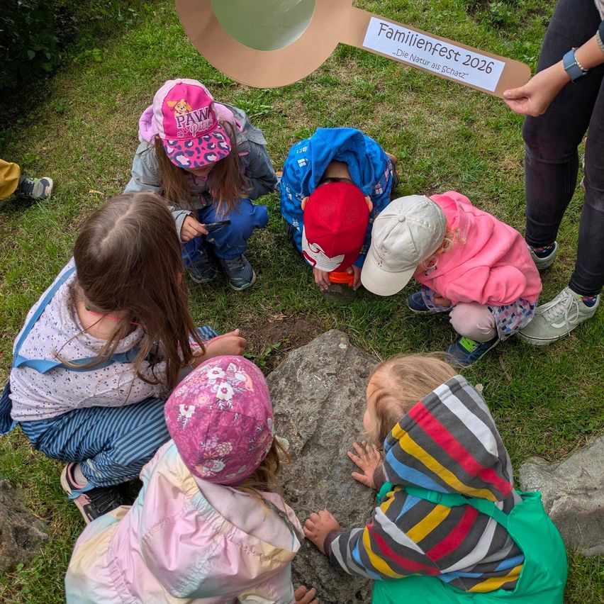 Eine Gruppe von Kindern und einer Lehrerin erforscht einen Felsen im Freien. Einige tragen Hüte, Turnschuhe und Jacken. In der Nähe steht ein Holzschild mit dem Text 'Familienfest 2026.'