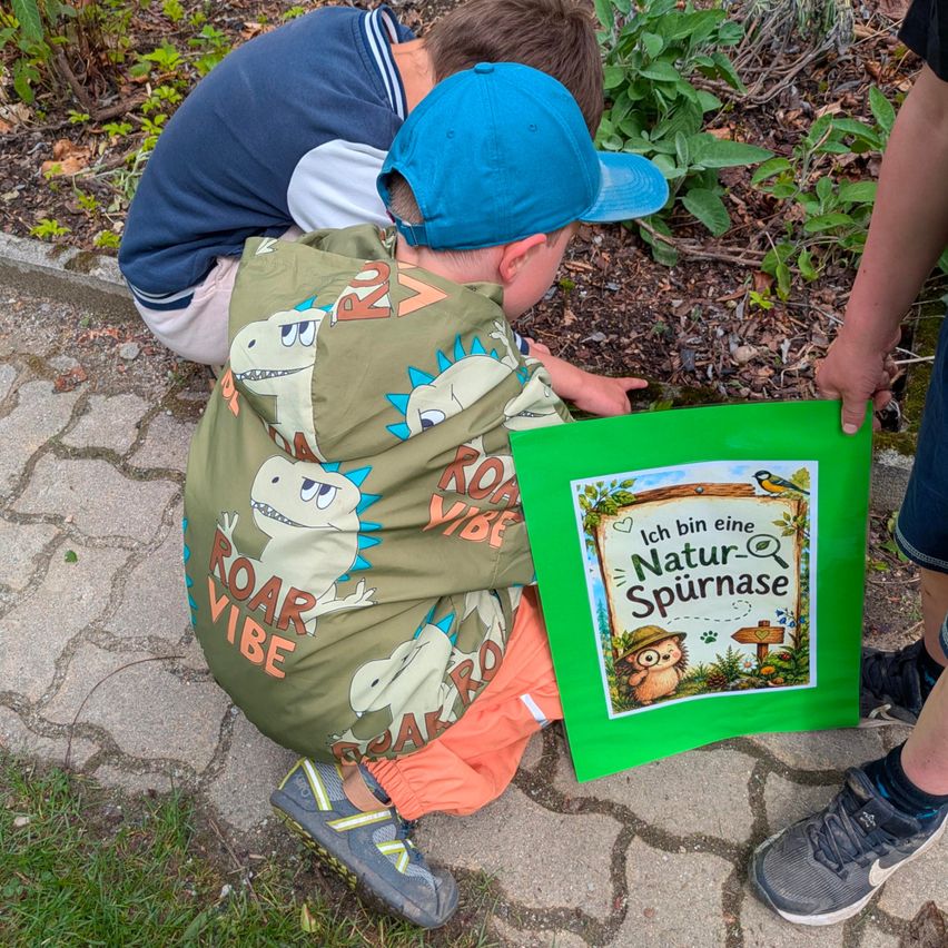 Zwei Jungen bücken sich, um Pflanzen in einem Garten zu betrachten. Einer hält einen grünen Papierbeutel mit dem Text 'Ich bin eine Natur-Spurnase.' Ein weiterer Junge hinter ihm trägt eine blaue Kappe.
