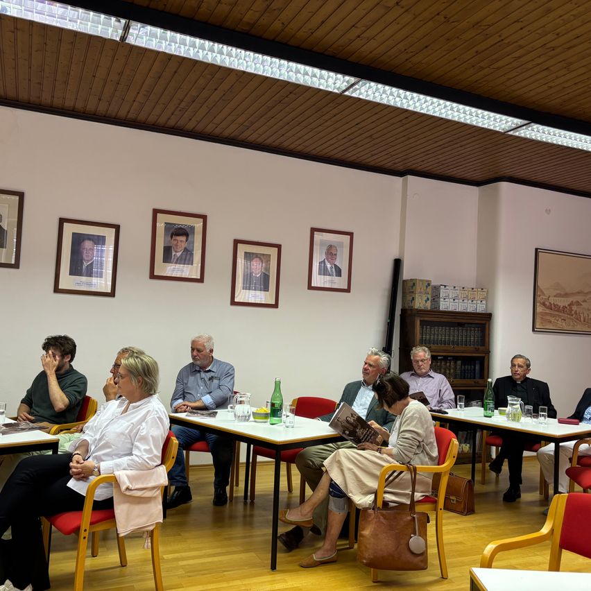 Eine Gruppe von Menschen sitzt um Tische in einem Raum mit gerahmten Porträts an der Wand. Sie scheinen sich in einer Besprechung oder Diskussion zu befinden.