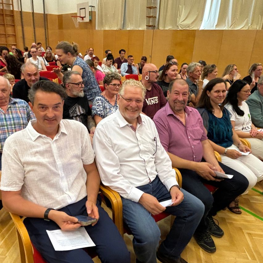 Eine Gruppe von Menschen sitzt auf Stühlen in einem Innenraum, möglicherweise einem Konferenzsaal, lächelt und interagiert. Papiere und Telefone sind in ihren Händen.
