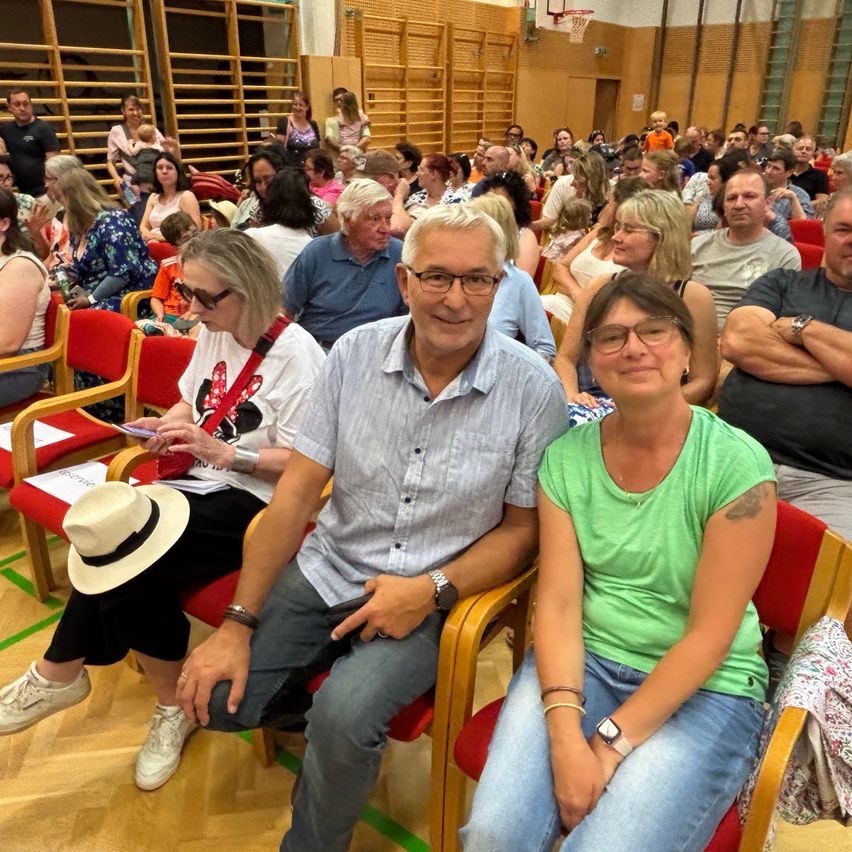 Eine Gruppe Erwachsener sitzt in Reihen roter Stühle in einem Saal, wahrscheinlich bei einer Versammlung. Einige tragen Brillen, Armbänder und Uhren. Ein Mann mit Brille hält ein Smartphone. Eine Frau neben ihm trägt ein Minnie-Mouse-Shirt.