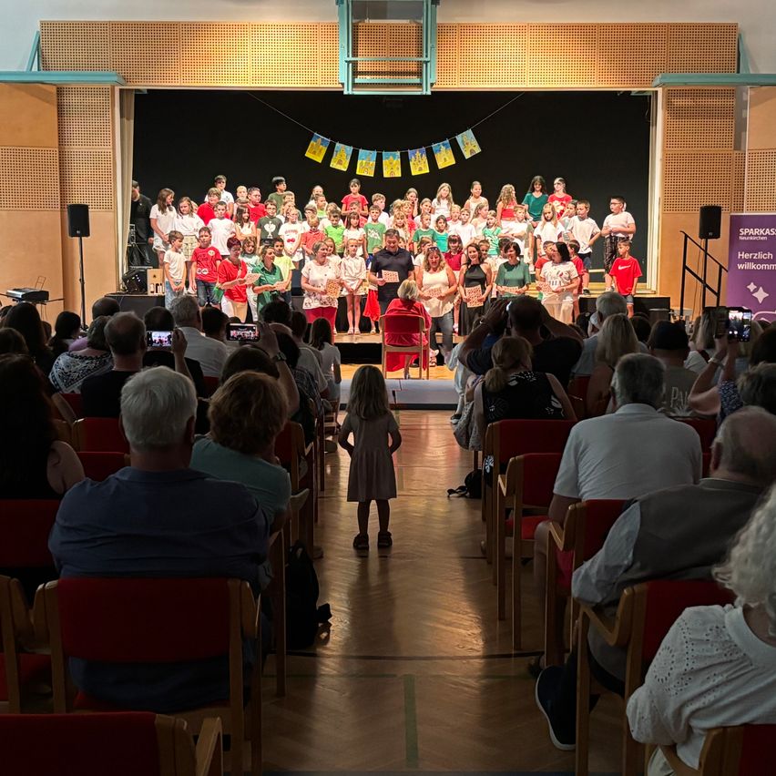 Eine große Gruppe von Kindern und Erwachsenen steht auf der Bühne, hält Papiere in der Hand und wird von einem Publikum in einem Saal beobachtet. Bunte Wimpel wehen über ihnen.