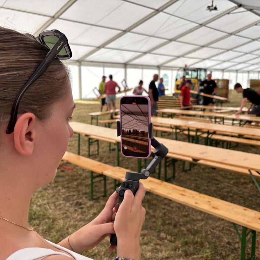 A woman takes a photo with her phone at an outdoor event, wearing sunglasses and a necklace. Behind her, several people are working under a tent.