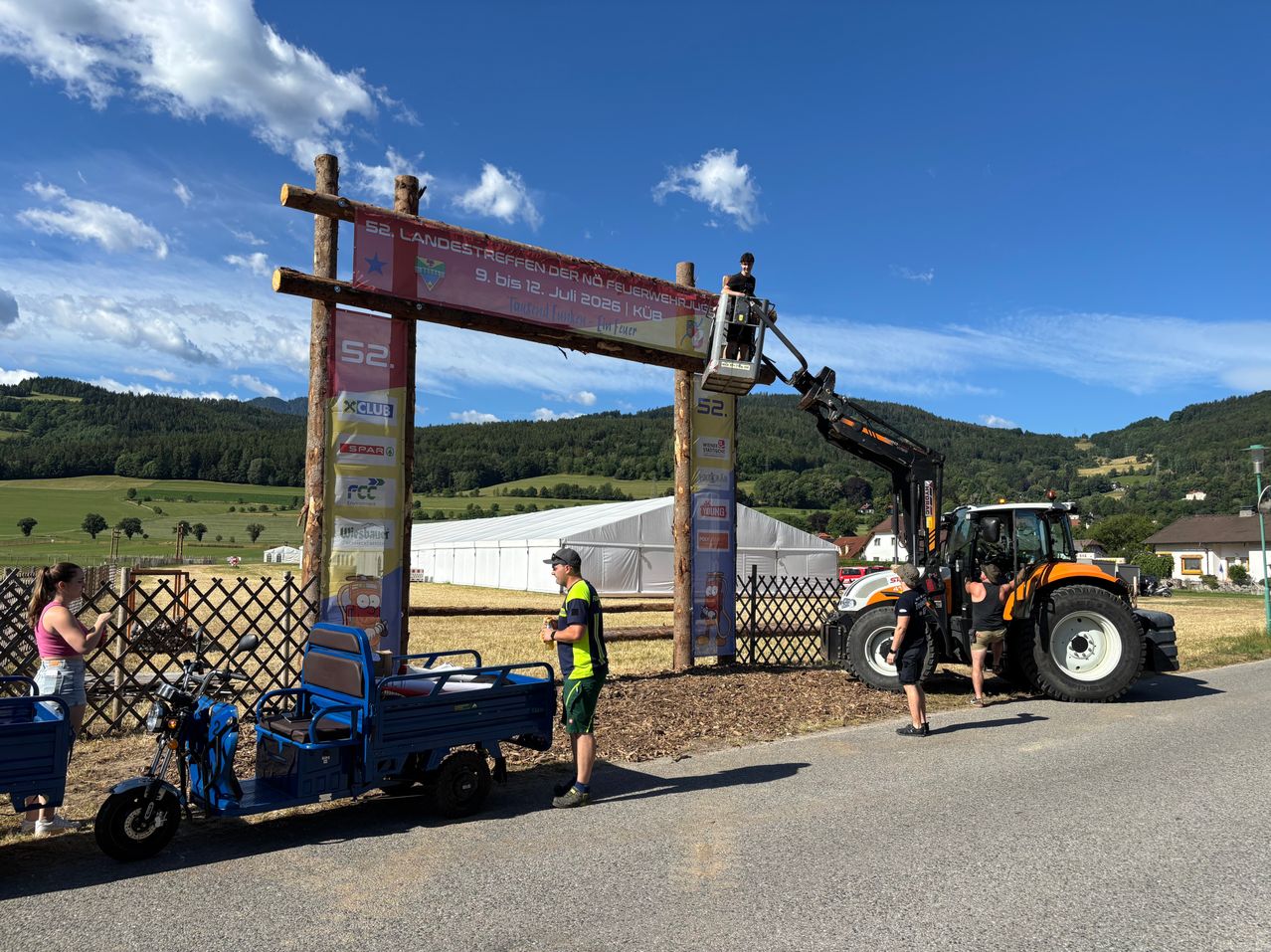 At an outdoor event, a person operates a crane to install a red banner above a wooden arch, while others stand nearby and a tractor is parked close by.