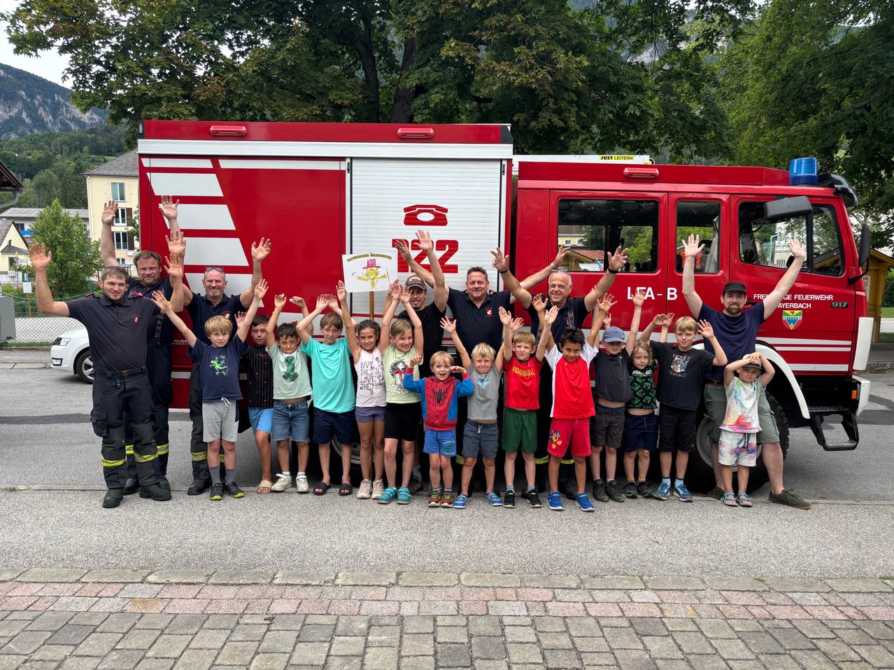 A group of firefighters and children pose in front of a red fire truck. Trees and buildings are in the background.