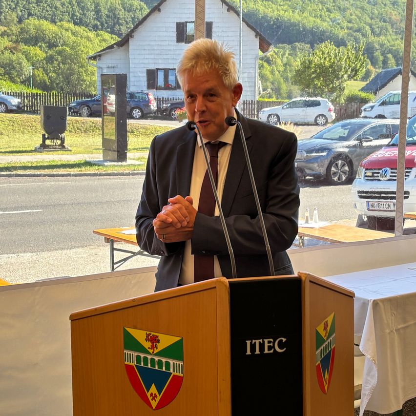 Ein Mann in einem Anzug spricht in ein Mikrofon an einem Podium mit einem Logo, umgeben von geparkten Autos und einem Haus im Hintergrund.