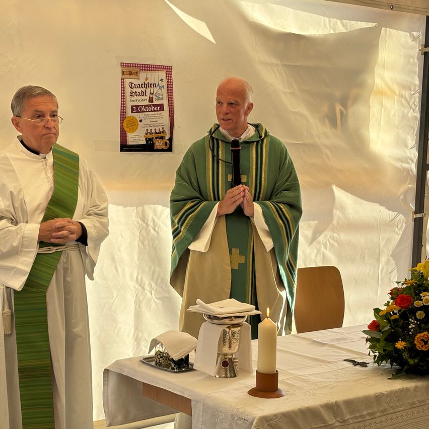Zwei Priester stehen in einem Zelt, einer hält ein Mikrofon, der andere betet. Vor ihnen steht ein Tisch mit einer Kerze und Blumen.