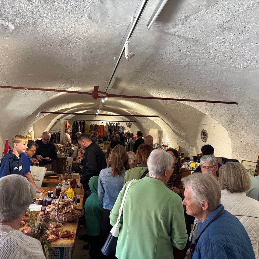 Eine Gruppe von Menschen versammelt sich in einem tunnelartigen Raum, wahrscheinlich auf einem Markt. Sie betrachten die ausgestellten Gegenstände auf den Tischen.