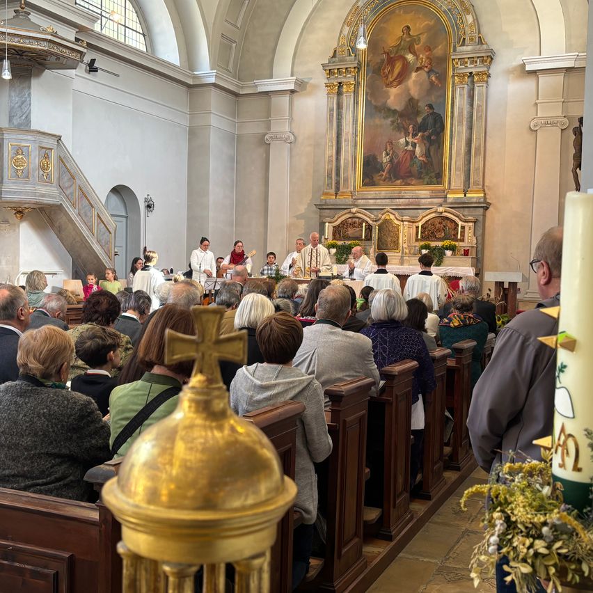 Eine Gemeinde von Menschen ist in einer Kirche versammelt und hört einem Priester am Altar zu. Der Altar ist mit einem Gemälde und Kerzen geschmückt. Die Kirche ist mit hölzernen Kirchenbänken gefüllt und ein goldenes Kreuz ist im Vordergrund sichtbar.