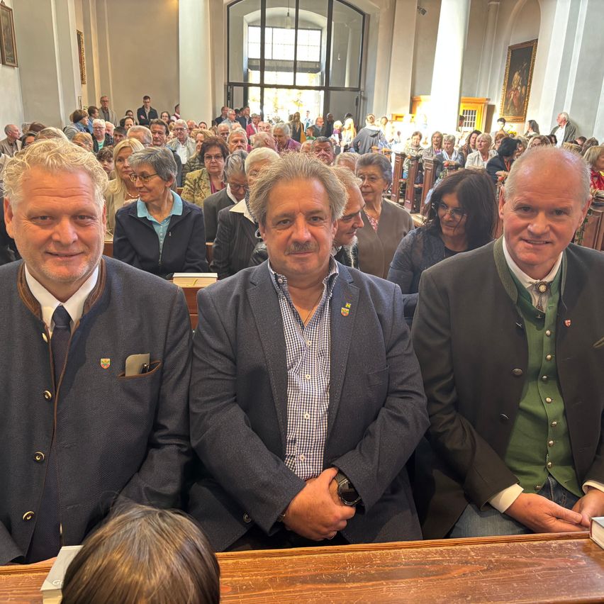 Drei Männer in Anzügen sitzen in einer Kirche mit einer Menge Menschen hinter ihnen.