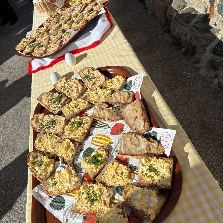 Ein Tisch mit einem gelben und weißen karierten Tischtuch ist draußen aufgestellt. Es gibt zwei Holzschalen mit verschiedenen Sandwiches, einige davon mit Eiern und Kräutern belegt.