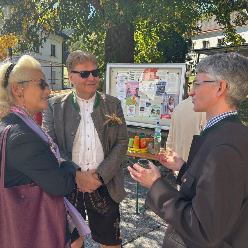 Drei Personen stehen vor einer Pinnwand mit verschiedenen Plakaten und einem Tisch mit Getränken.