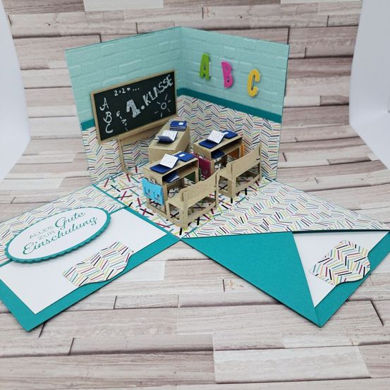 Eine Miniatur-Klassenzimmerszene mit Tafel, Tischen und Stühlen, auf einem hölzernen Hintergrund mit einem 'Gutes fur Einschulung'-Etikett.