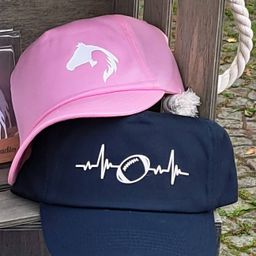 Zwei Mützen auf einer Holzoberfläche gestapelt; eine rosa Mütze mit einem weißen Pferdedesign und eine marineblaue Mütze mit einer Herzfrequenzlinie und einem Football-Symbol.