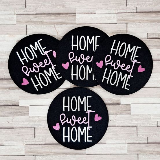 Vier runde Matten mit den Worten 'Home Sweet Home' in Weiß und Rosa auf schwarzem Hintergrund, auf einer Holzoberfläche angeordnet.