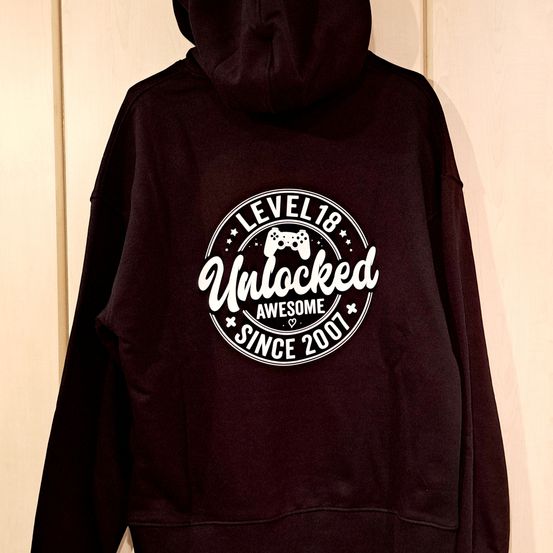 Ein schwarzer Hoodie mit einem weißen kreisförmigen Design, das 'LEVEL 18 Unlocked AWESOME Since 2001' mit einem Game-Controller-Symbol im Inneren des Kreises liest.
