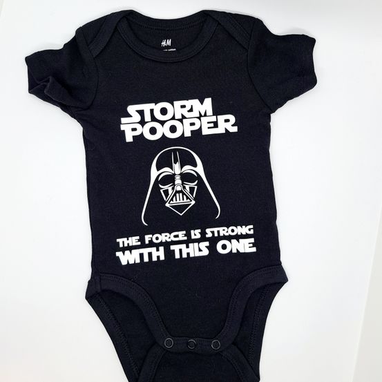 Ein schwarzes Babybodysuit mit weißem Druck, der 'Storm Pooper' und ein Darth Vader Logo zeigt, zusammen mit dem Zitat 'Die Macht ist stark mit diesem'.