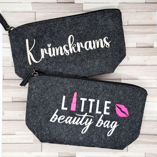 Zwei graue Filzstifttaschen mit 'Krimskrams' und 'Little beauty bag' bedruckt. Die erste Tasche hat ein rosa Lippenstift-Symbol und die zweite hat ein rosa Herz-Symbol.
