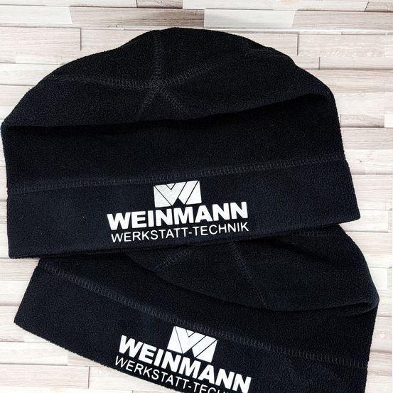 Zwei schwarze Mützen mit weißen Logos, die 'WEINMANN WERKSTATT-TECHNIK' lesen, auf einer Holzoberfläche gestapelt.