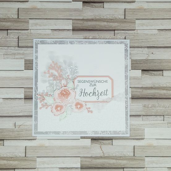 Eine Hochzeitskarte mit einem Kreuz, rosa Blumen und dem Text 'Segenwunsche zur Hochzeit' wird auf einem hölzernen Hintergrund gezeigt.
