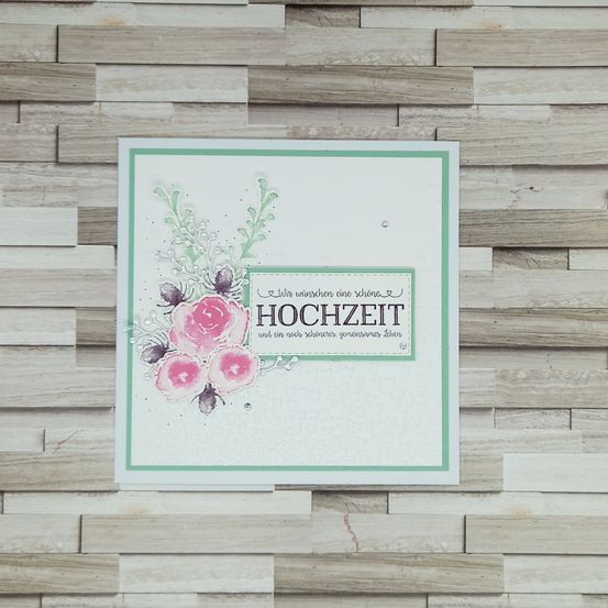 Eine Grußkarte mit einem Blumendesign, mit rosa Blumen und grünen Blättern, auf einem hölzernen Hintergrund. Die Karte trägt 'Hochzeit' in fetten Buchstaben, mit einem Zitat darunter.