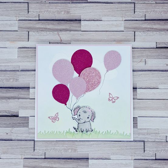Eine handgefertigte Karte mit einem niedlichen Elefanten, der pinke Glitzerballons und Schmetterlinge auf einem hölzernen Hintergrund hält.