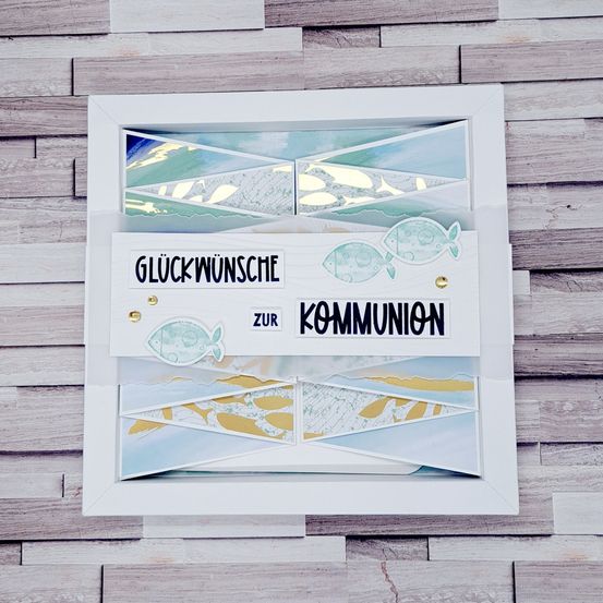 Eine weiße Karte mit einem Fischdesign und den Worten 'Gluckwunsche zur Kommunion' in Blau. Die Karte liegt auf einer Holzoberfläche.