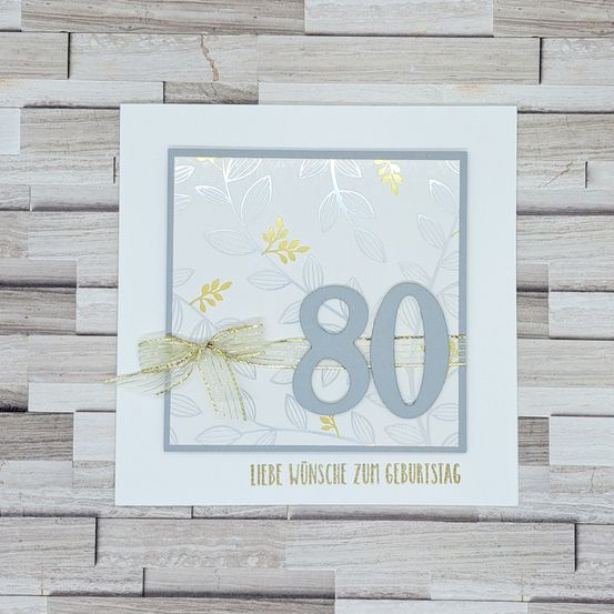 Eine Geburtstagskarte mit der Nummer 80 in Silber und Gold, begleitet von einer goldenen Schleife und einem Blattmuster auf einem hölzernen Hintergrund.