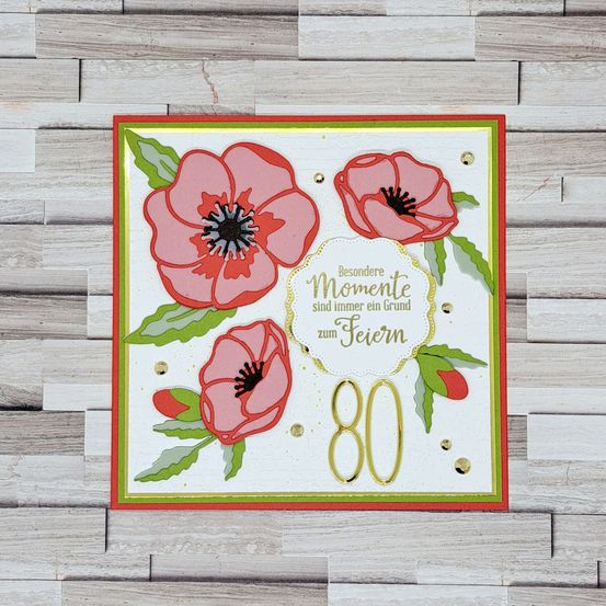Eine Grußkarte mit einem roten Mohn-Design und der Nummer 80 in Goldfolie, zusammen mit dem Text 'Besondere Momente sind immer ein Grund zum Feiern'. Die Karte liegt auf einem hölzernen Hintergrund.