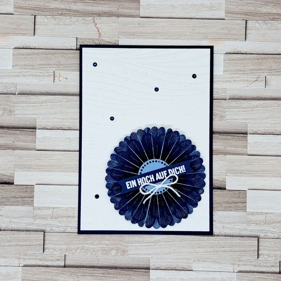 Eine dekorative Karte mit einem blauen Blumendesign und einem darum gebundenen Band, mit der deutschen Phrase 'EIN HOCH AUF DICH!' auf einem hölzernen Hintergrund.
