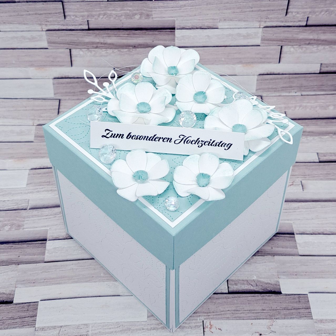 Eine blaue und weiße Schachtel mit weißen Blumen und einem Etikett, das Zum besonderen Hochzeitstag steht. Die Schachtel steht auf einer hölzernen Oberfläche.