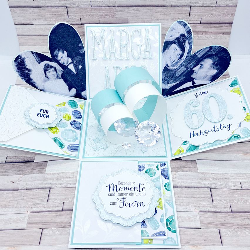 Eine Pop-up-Karte mit blau-weißem Design zur Feier eines 60. Jahrestages. Sie zeigt eine Fotocollage eines Paares, die Worte 'Marga' und 'Hochzeitstag' und einen Kristall als Mittelpunkt. Die Karte ist in Kreuzform gefaltet und auf einer Holzoberfläche platziert.