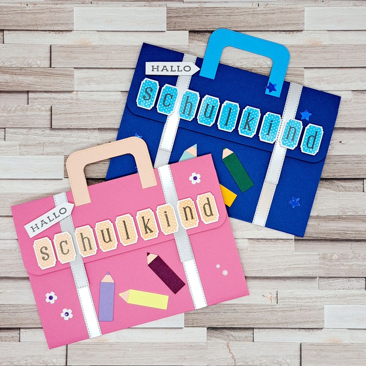 Zwei bunte Papiertüten mit 'Hallo Schulkind' darauf, eine pink und eine blau, auf einem hölzernen Hintergrund mit Stiften und Blumen.